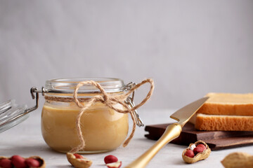 Glass jar with natural peanut butter, golden colored knife, peanuts and toast sandwiches on a light background. Vegetarian traditional breakfast close up. Copy space