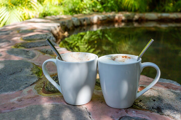 Two white ceramic mugs, one with cappuccino coffee and the other empty with metal spoons on the edge of an outdoor stone Jacuzzi reflecting the plants around it, sunny spring day