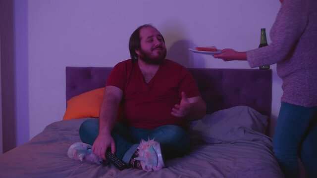 Young fat guy watching tv in bed while senior mother bringing him beer and sandwich