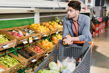 Young man standing near shopping cart and fresh fruits on counter in store