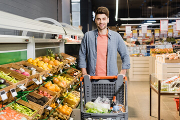 Smiling man looking at camera near shopping cart in supermarket