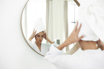 Obraz premium Young woman in bathrobe looking in bathroom mirror. Pure beauty. Attractive woman touching her face and smiling while looking in the mirror