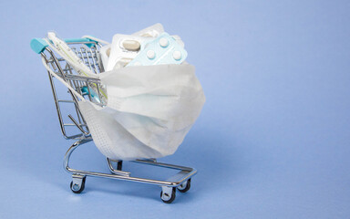 Shopping cart and medicine. An expensive medicine. Cough pills shopping basket with thermometer and medicine mask on blue background.
