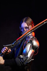 Futuristic female fighter with glowing sword and eyewear in black background. Attractive military woman looks at camera hiding her face behind collar.
