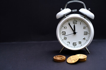 classic white alarm clock stands next to some gold coins and the time shows 5 minutes to 12