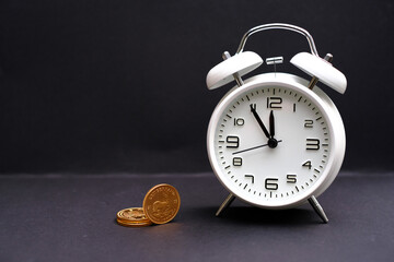 classic white alarm clock stands next to some gold coins and the time shows 5 minutes to 12