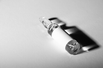 Black and white photo. Selective focus ampoule with medicine with copy space. The vaccine vial lies on white surface and casts dark shadow. Pandemic concept. Health protection.