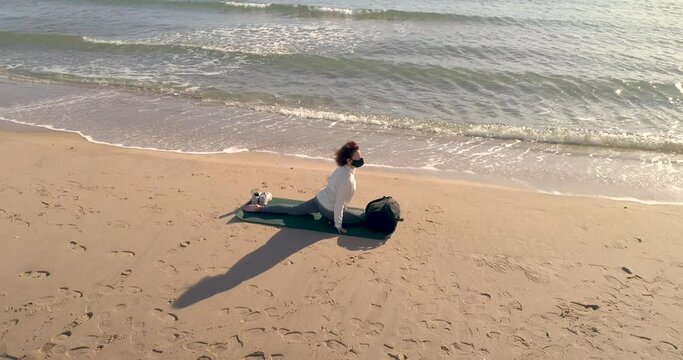 Mujer con mascarilla covid-19 realizando postura yoga en la arena de la playa y dron volando orbital