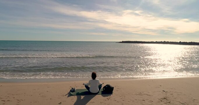 Mujer madura sentada mirando al mar sentada en la arena en postura de meditaci&oacute;n