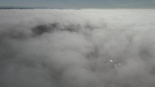 Drone footage above the clouds, showing expanse of white soft fluffy clouds, with mountains in the distance, USA below, scattered clouds with peaks to the ground.