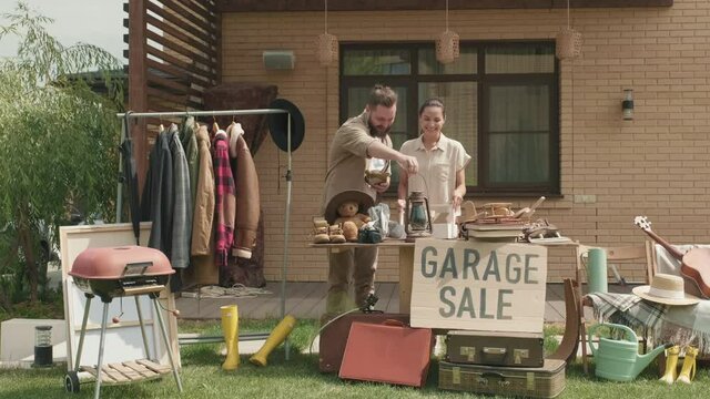 Handheld wide shot of cheerful man and woman chatting and preparing for garage sale in front yard of their suburban house in summer, then looking at camera and smiling