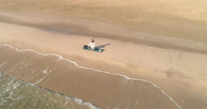 dron orbitando a mujer de mediana edad haciendo yoga sentada en la orilla del mar