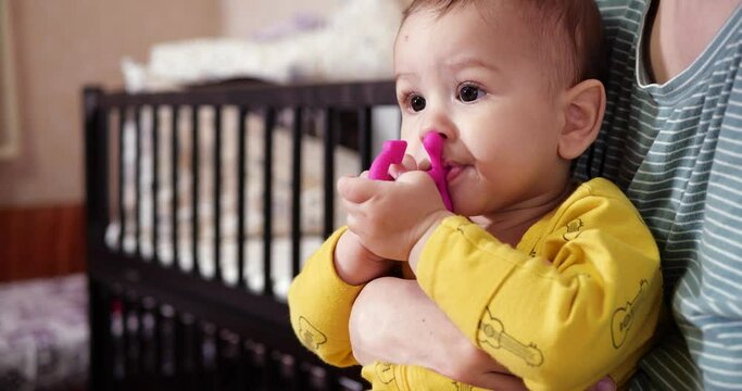 A young mother sits on the bed with a small child in her arms and controls the game with a teether and other tactile toys. Maternity, motherhood and family concept