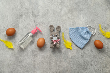 Fototapeta na wymiar Creative festive flat lay. Eggs, protective face mask, antiseptic, and handmade knitted rabbit on gray background. Happy Easter during the coronavirus pandemic. Top view, copy space