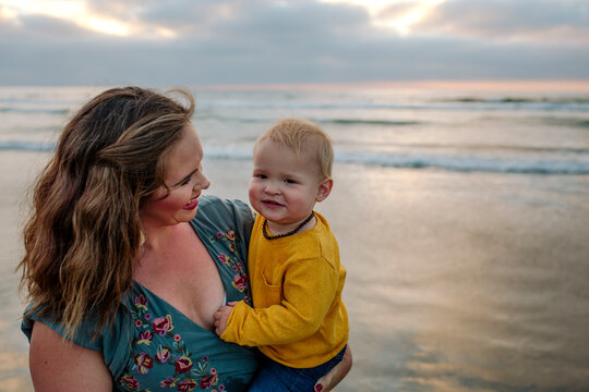 Beautiful young mother holds smiling 6 mo old baby at ocean at sunset
