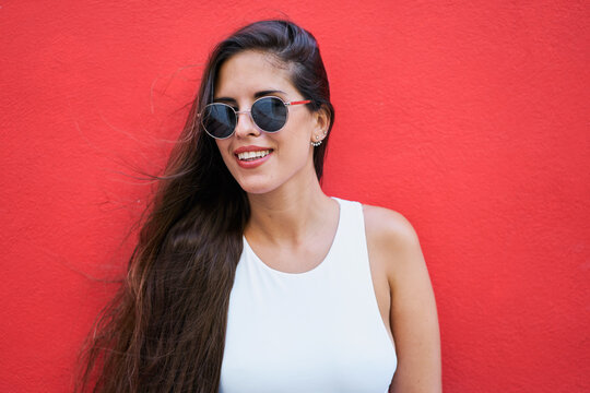 Young woman with long hair wearing stylish sunglasses and standing near red building wall in city
