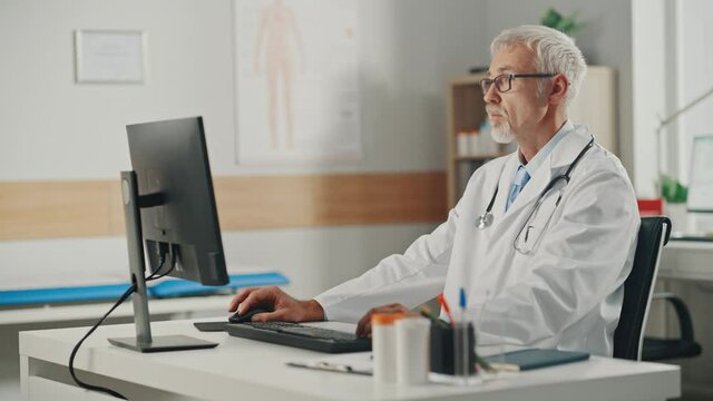 Experienced Middle Aged Male Doctor Wearing White Coat Working on Personal Computer at His Office. Senior Medical Health Care Professional Working with Test Results, Patient Treatment Planning.