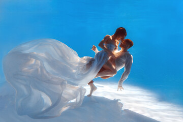 The couple is dancing or hugging in the pool underwater. A girl in a dress with a long train and a guy swim underwater