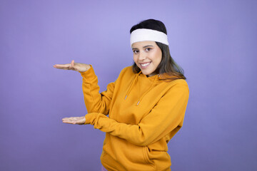 Young latin woman wearing sportswear over purple background gesturing with hands showing big and large size sign, measure symbol. Smiling . Measuring concept.