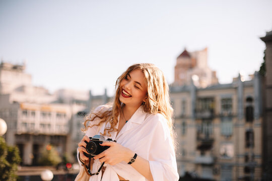 Happy young woman holding camera while standing in city during summer