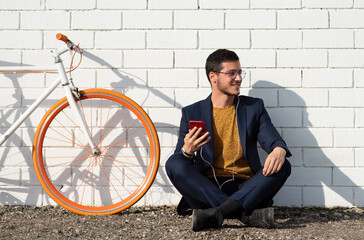 Obraz na płótnie Canvas Hobre joven de negocios consultando su celular que se dirige al trabajo en bicicleta vintage blanca y apoyado en pared blanca de ladrillos.