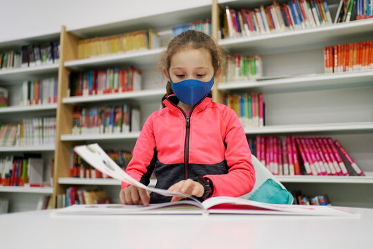 Little girl wearing a medical mask and reading a book in a libra