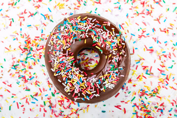 Top view of a chocolate donut covered with many colorful sprinkles on a white table full of sweet sprinkles