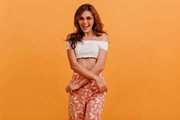 Emotional positive lady in orange pants crossed her arms and poses for portrait, laughing and having fun in studio