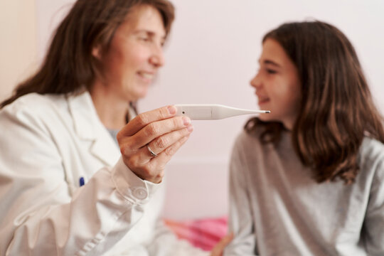 Female doctor teaching the temperature on a thermometer to a girl in her bed. Home doctor concept