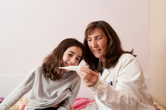 Female doctor teaching the temperature on a thermometer to a girl in her bed. Home doctor concept