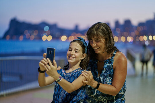 Portrait of mother and teenage daughter wearing blue dresses and taking a selfie with their smartphone at night with a beach in the background. Vacation concept