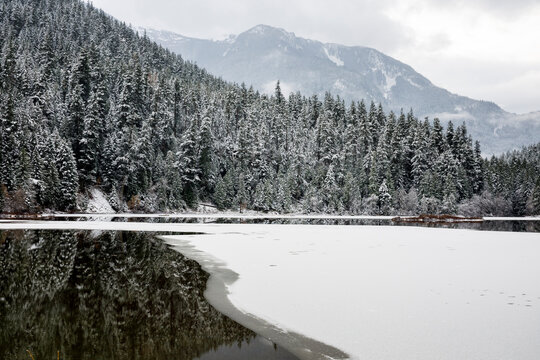 Lake surrounded by mountains and snow covered forest starts to freeze