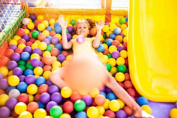 Fototapeta na wymiar A funny girl sits in a playground with soft and bright equipment and throws colorful balls towards the camera while enjoying the warm summer sun