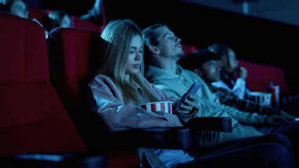 Beautiful young woman using her phone for texting while missing boring movie at the cinema