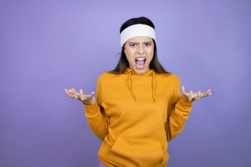 Young latin woman wearing sportswear over purple background crazy and mad shouting and yelling with aggressive expression and arms raised. Frustration concept.