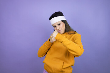 Young latin woman wearing sportswear over purple background Punching fist to fight, aggressive and angry attack, threat and violence