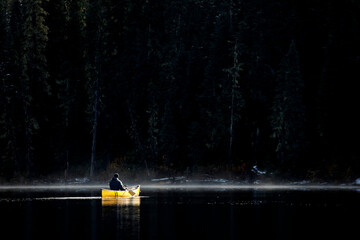 Rugged man solo paddles canoe on misty calm lake by thick forest