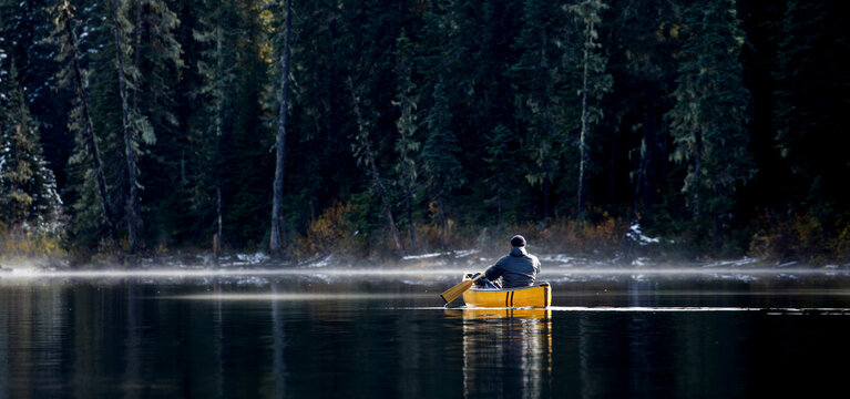 Man paddles canoe on lake with mist and fog on sunny day by forest