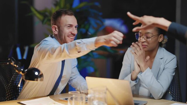 Cooperative team of multi-ethnic coworkers organise business meeting sitting together and telling jokes. Friendship company. Night office concept.