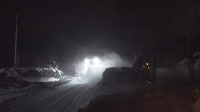 Back view on tractor Cleaning and blowing Snow at Night in A Snowfall at countryside road, heavy wind blows snow flakes across whole road