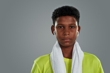 Active lifestyle. Close up portrait of a confident teenage african boy in neon shirt with white towel on shoulders wearing wireless earphones looking at camer