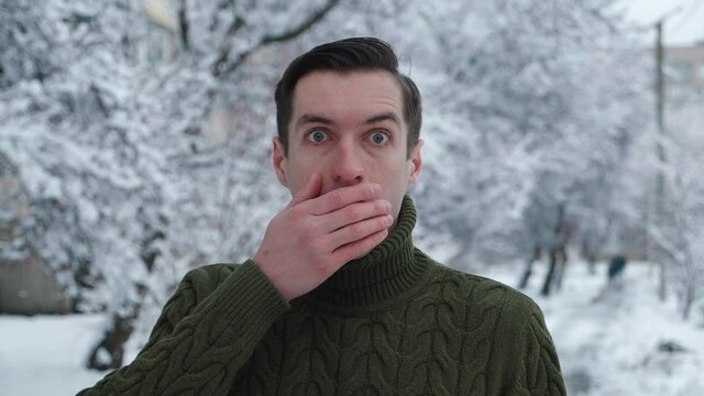 Shocked young male saw something terrible, fearing for his life outdoor at wintertime. Portrait of frightened young man closes his mouth with hand outdoors at winter snowy background
