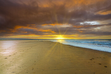 Deserted beach during sunset with orange deep blue colored clouds and low sun with sun rays on the horizon shining over the beach