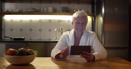 Senior man wearing bathrobe using digital tablet sitting at table in kitchen late at night - Powered by Adobe