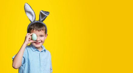 Happy child boy with bunny ears is covering his eye with Easter egg on yellow background. A kid with Easter egg on yellow background. Copy space