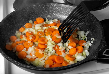 Vegetables are fried in a pan on the stove. Finely chopped onions and carrots are stewed in a pan. The food is cooked in the home kitchen. 