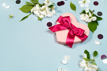 Heart shape pink present box with red ribbon bow, blooming white flowers border on blue background with copy space. Beautiful blossom banner template. Spring birthday, valentine day love concept