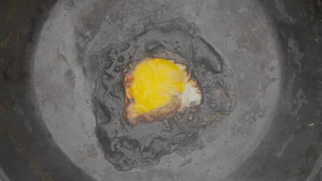 Overhead shot: an egg forgotten in a rusty pan, its albumen completely turned black, cooking for too long, releasing an ominous smoke.
