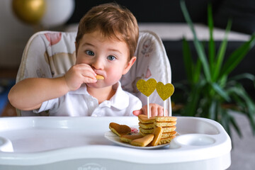 little boy sits on a baby chair and eats heart-shaped pancakes. Happy valentine's day. Funny...