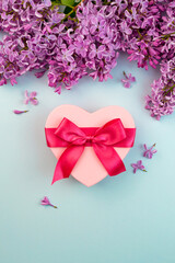 Heart shape pink present box with red ribbon bow, Lilac branch flower blooming bouquet border on blue background. Spring birthday, 14 february love still life concept. Beautiful greenery blossom
